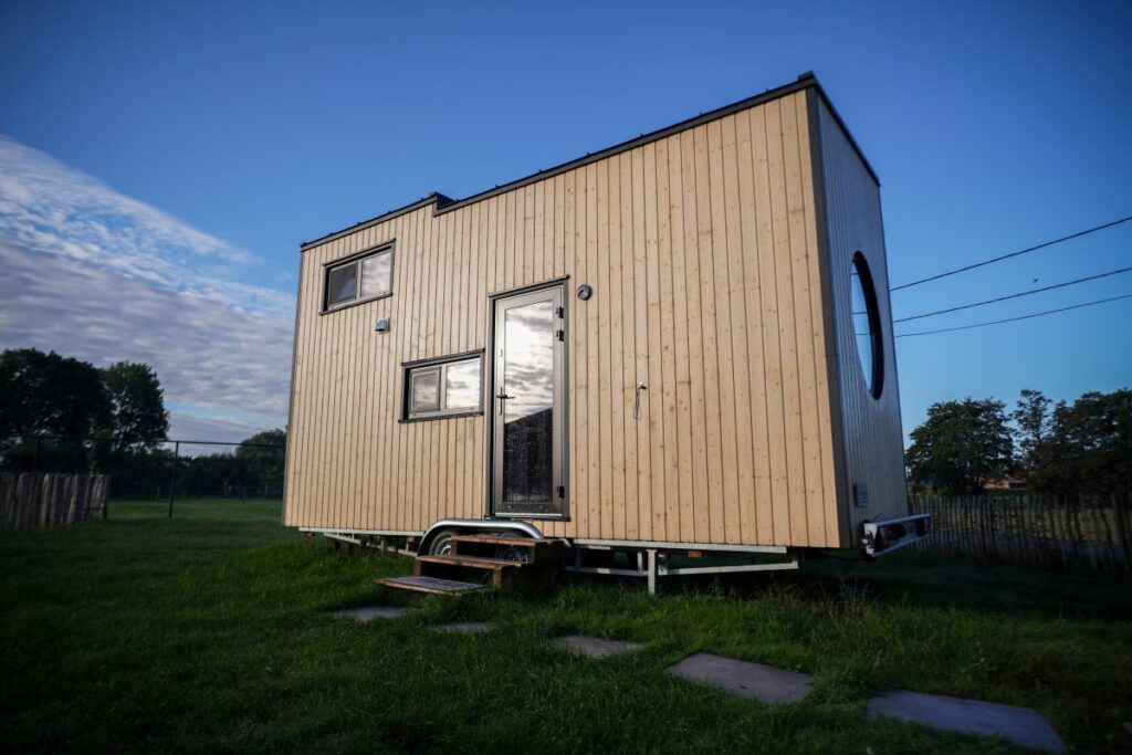 Een modern huisje met een licht houten buitenkant staat op een grasveld onder een blauwe hemel, met meerdere rechthoekige ramen en een rond raam aan de zijkant. Trappen leiden naar de glazen deur aan de voorkant.