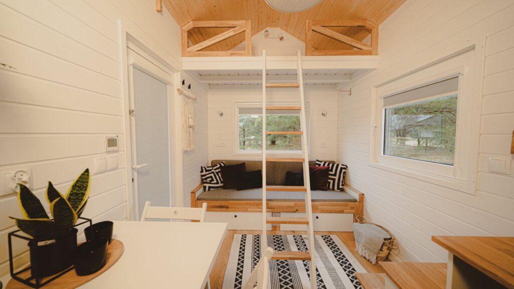 Licht, gezellig tiny house interieur met witte muren, een houten vide bereikbaar via een ladder, een kleine bank met zwart witte kussens, grote ramen, een tafel met stoelen en potplanten op een rond dienblad.