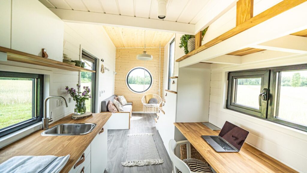 Licht, modern tiny house interieur met houtaccenten, met een keuken aan de linkerkant, een werkruimte met een laptop aan de rechterkant en een gezellige zithoek met een rond raam aan het uiteinde, met uitzicht op groene velden.