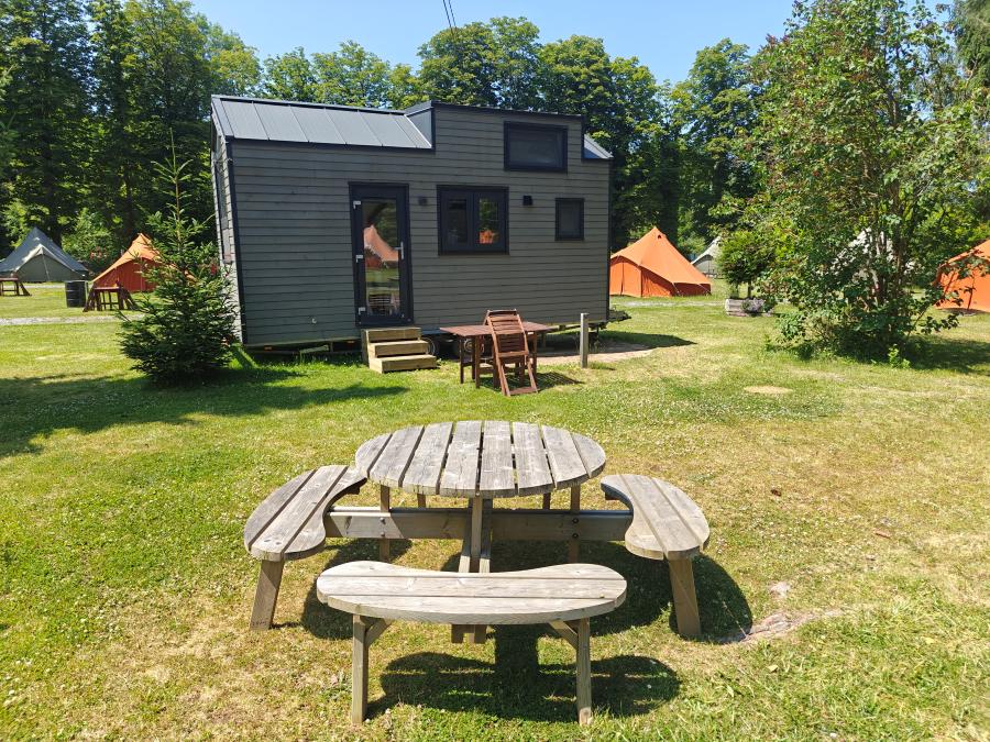 Een klein grijs huisje met een zwart dak staat op een grasveld met een houten picknicktafel en banken ervoor. Op de achtergrond zijn oranje tenten en groene bomen te zien onder een zonnige hemel.