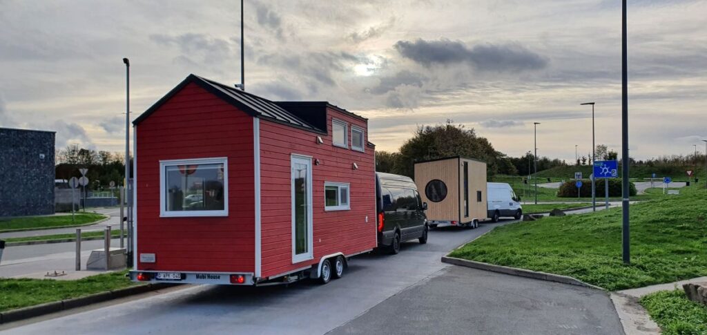 Twee voertuigen slepen kleine huisjes langs een weg onder een bewolkte hemel. Het eerste huisje is rood met witte bies; het tweede is beige met een rond raam. Straatverlichting en gras omzomen de weg.