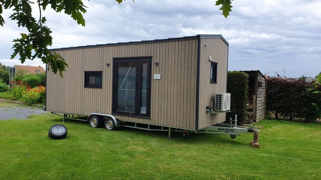 Een modern huisje op wielen met houten panelen, grote glazen deuren en kleine ramen staat geparkeerd op een grasveld. Vlakbij ligt een los reservewiel en aan de zijkant is een airconditioning gemonteerd.
